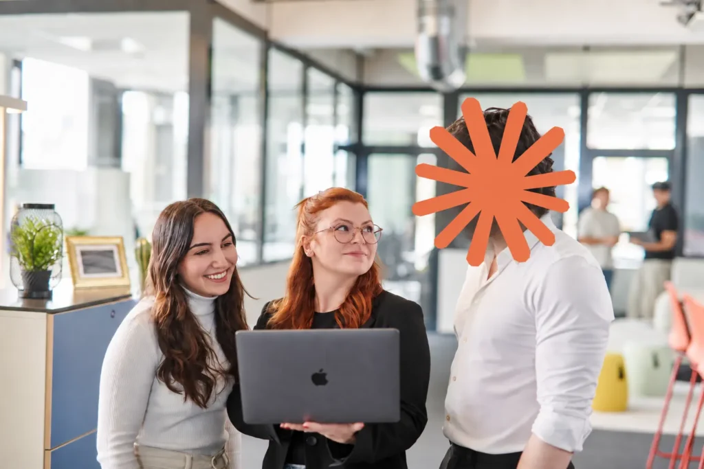 Zwei Kolleginnen mit Laptop stehen im hellen Büro bei einem Kollegen, dessen Gesicht ein orangefarbenes Claude-Logo ist.